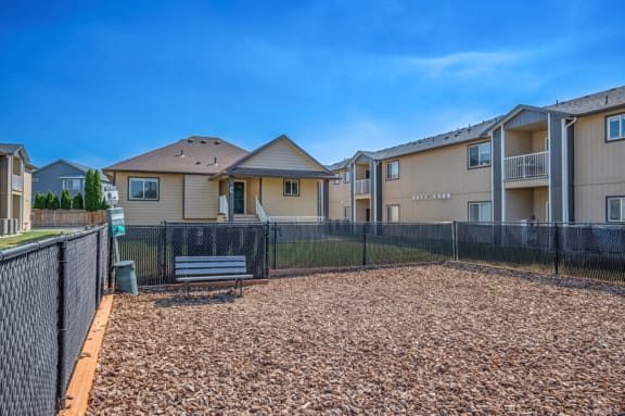 Fenced in dog park with bench and bag dispensing station. Surrounded by green grass and apartments.at Pine Tree Park, Washington, 99337