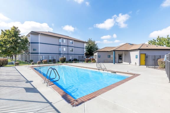 A bright blue swimming pool with lounge chairs, picnic tables and a clubhouse to the right.at Shoreline Village, Washington, 99352