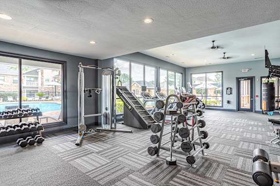A spacious gym with a variety of equipment and a view of a pool and houses outside the windows at Avenues at Shadow Creek Ranch Apartments, Pearland