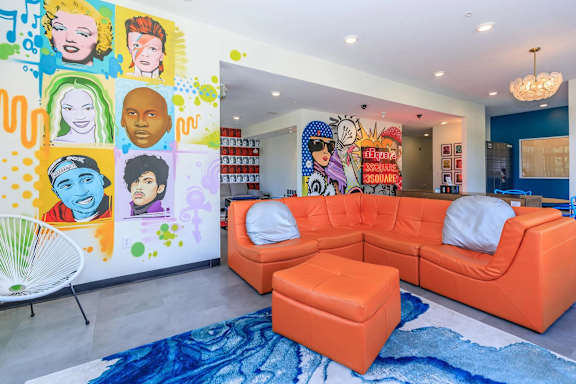 A brightly colored living room with a large orange sofa and a mural of various faces on the wall.