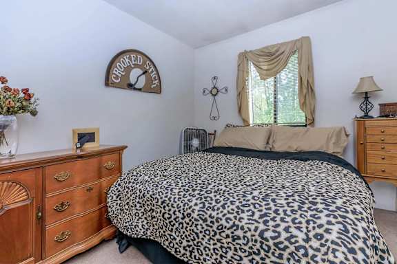 A leopard print bed sits in a bedroom with a wooden dresser and a cross hanging on the wall.