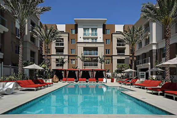 A large swimming pool in front of a multi-story apartment building with red lounge chairs on the side.