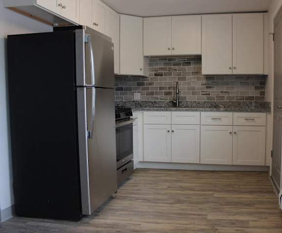 A black refrigerator stands in a kitchen with white cabinets and a tiled backsplash.