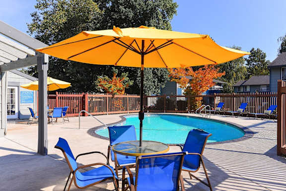 A yellow umbrella shades a table with blue chairs around a pool. at The Lakes at Fife, Fife, WA, 98424