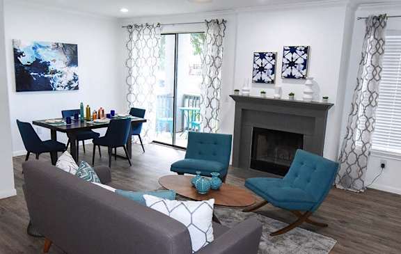 A living room with a grey couch, a blue chair, a grey armchair, a grey coffee table, a grey fireplace, a grey dining table with blue chairs, a painting, and a painting on the wall.