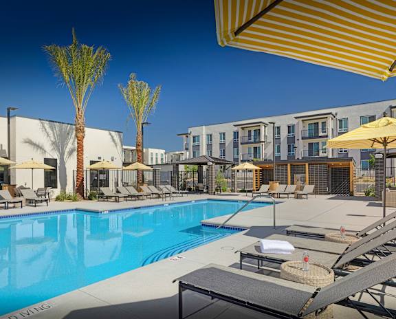 A pool area with sun loungers and umbrellas in front of apartment buildings.