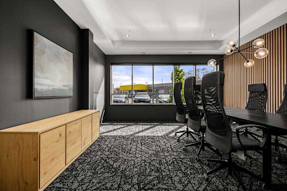 A room with a wooden cabinet, a painting, a window with a view of the outside, a carpet, a table with chairs and a lighting fixture.