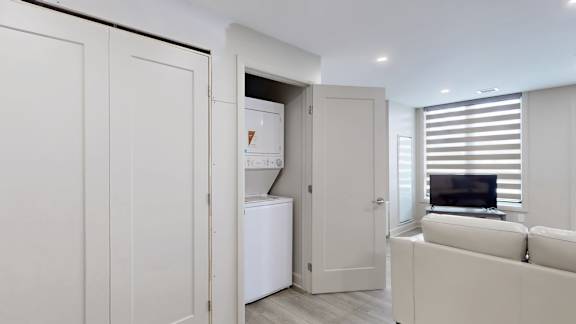 A white laundry room with a washer and dryer is open to a living room.