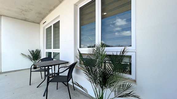 A table and chairs are on a balcony with a window and plants.