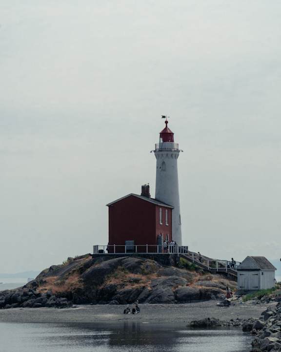 A red and white lighthouse stands on a rocky outcrop by the water.