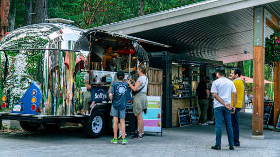 A food truck with a silver exterior and a blue and white logo on the side is parked outside a building with people standing in front of it.