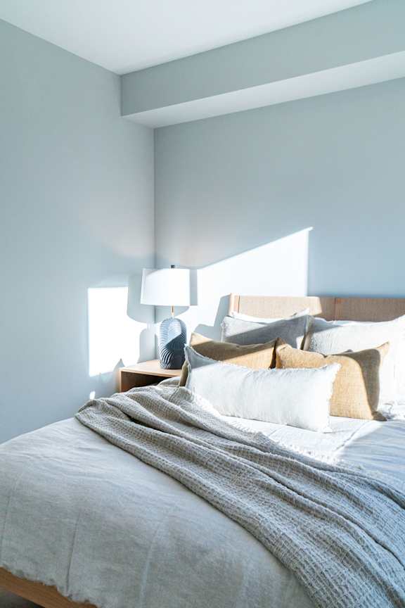A bed with a grey blanket and pillows is in the foreground with a lamp on a side table in the background.