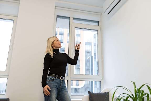 A woman in a black top and blue jeans is standing in a room with a window.