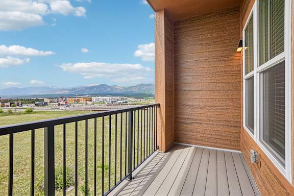 A balcony with a black railing and a wooden floor overlooks a grassy field and distant mountains.