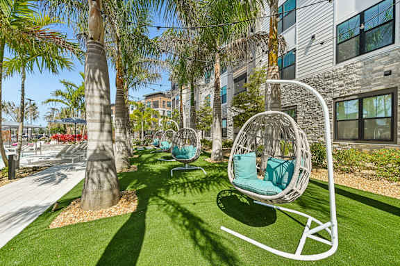 A white swing chair with a green cushion is in the foreground of a sunny outdoor area with palm trees and a building in the background at The Concord Luxury Apartments, Florida