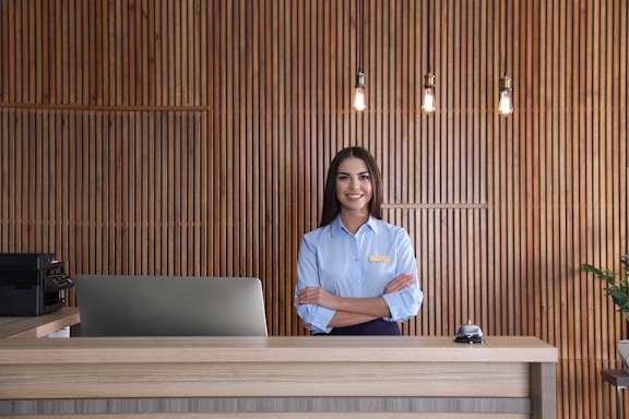 A woman in a blue shirt is standing in front of a wooden wall with a laptop and a plant on the desk.