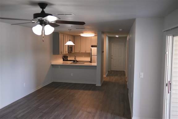 A room with a ceiling fan and a kitchen area in the background.