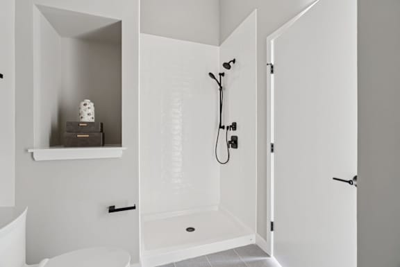 A white bathroom with a toilet, a shelf with two objects on it, and a shower with a hose.