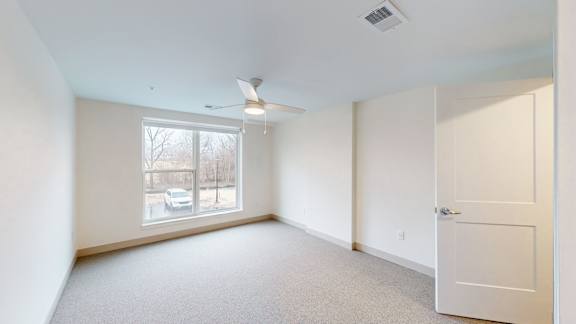 A room with a white door, carpeted floor, and a window showing a view of a car and trees outside.