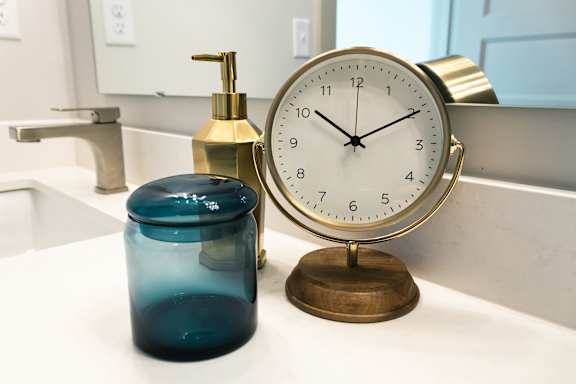 A blue jar sits next to a brass clock on a bathroom counter.