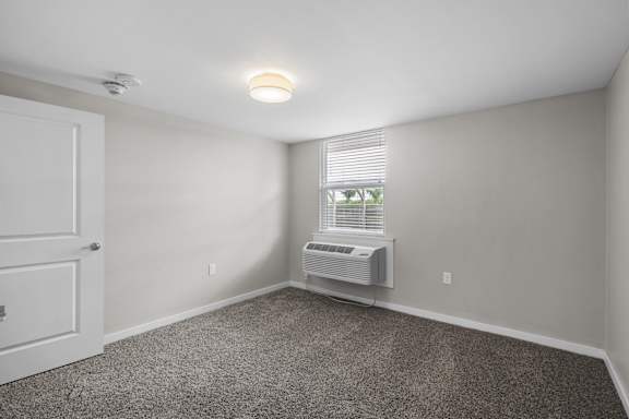 A room with a carpeted floor, a window with blinds, and a wall-mounted air conditioning unit.