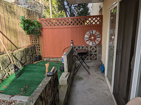 A small backyard with a green artificial grass area, a wooden fence, and a red wall with a clock.
