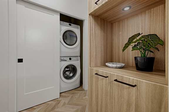 A white washing machine is in a small laundry room with a wooden cabinet and a plant.