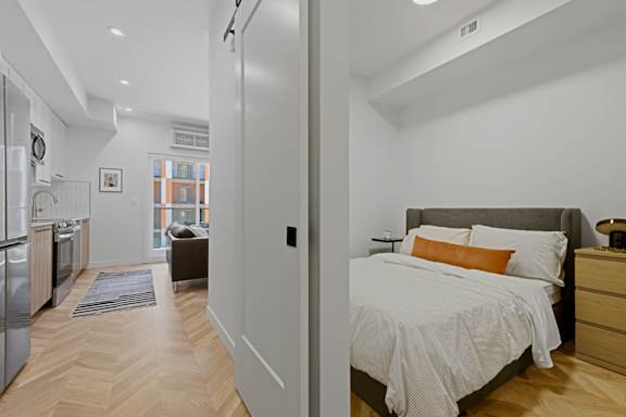 A bedroom with a bed, nightstand, and a kitchen area.