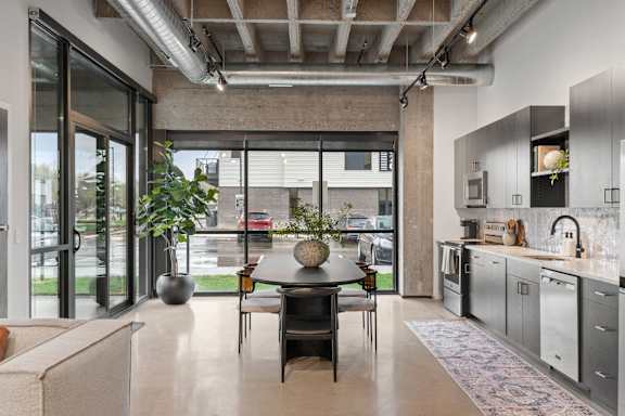 A modern kitchen with a dining table and chairs in the foreground and a view of the outside through the glass doors.