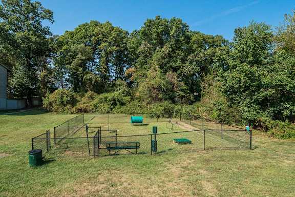A green trash can sits in a grassy field next to a tennis court.