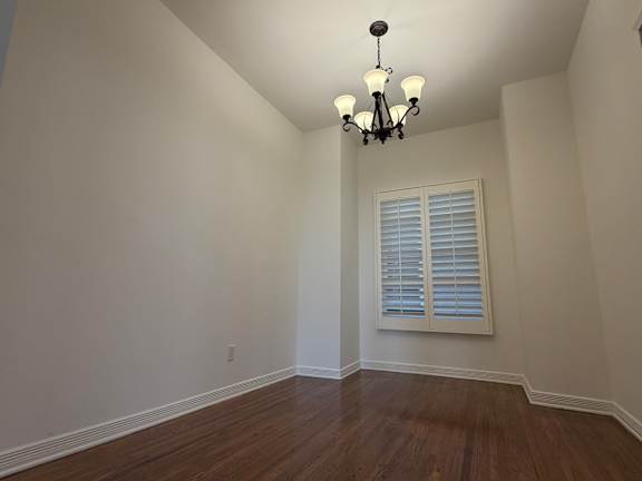 A room with a chandelier, a window with blinds, and wooden flooring.