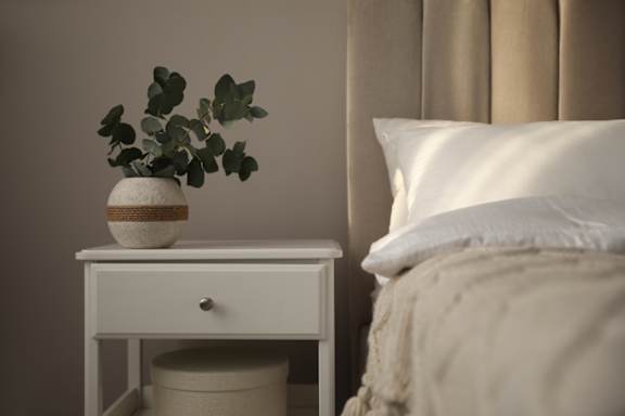 A white table with a plant on it sits next to a bed.