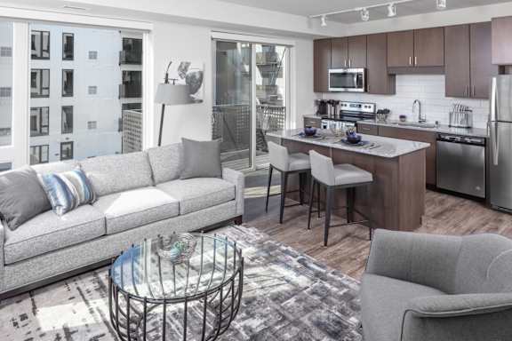 A modern living room with a grey sofa, a glass coffee table, and a kitchen area in the background.