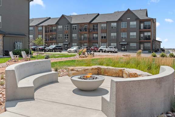 A modern outdoor seating area with a fire pit and concrete furniture in front of a multi-story apartment building.