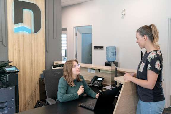 A woman is standing at a reception desk talking to another woman who is seated.