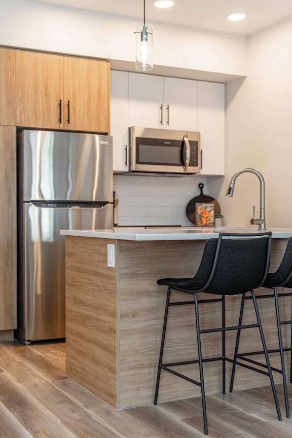 A kitchen with a stainless steel refrigerator, a microwave above the counter, and two black barstools.at Vibe Apartments at Arbour Lake, Calgary Alberta