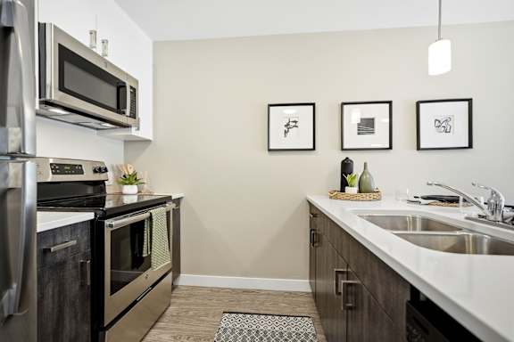 A kitchen with a stainless steel refrigerator and oven, a microwave above the stove, and a sink with a basket of fruit on the counter.