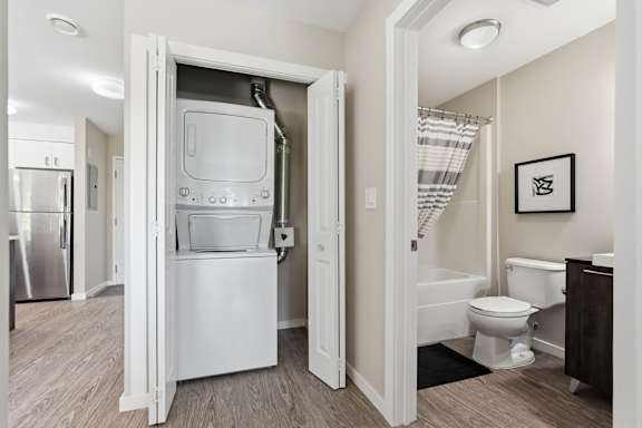 A white refrigerator is in the middle of a room with a toilet and a bathtub in the background.