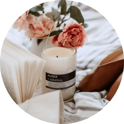 A candle labeled "AMBER MINERAL" sits on a bed with a white pillow and a white candle holder.