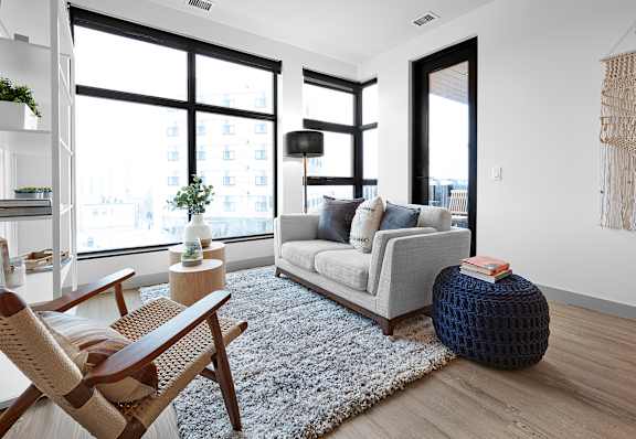 A living room with a grey couch, a wooden chair, a white rug, and a large window.at Raymond Block, Alberta