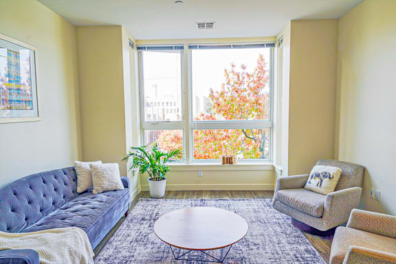 A living room with a blue couch, a grey armchair, a round table, and a window with a view of trees.