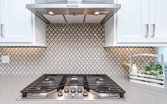 Gas range stove with five gas ranges sits in a beige marble countertop. There is an off-white tile backsplash on the wall below the white cabinets and stainless steel fume hood.