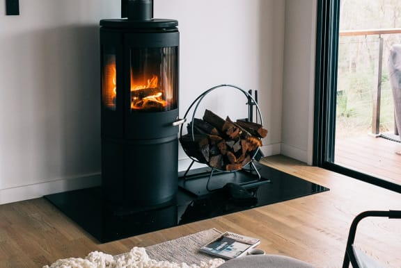 Wood-burning cylindrical stove-pipe fireplace in living room next to sliding glass balcony door. The fireplace is against a wall in a living room with light hardwood floors.