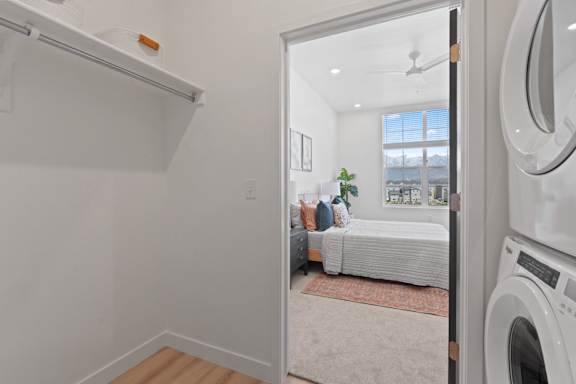 A white laundry machine is in the foreground of a room with a bed and a window.