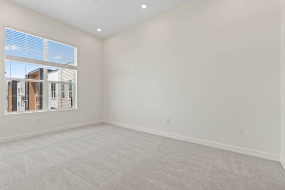 A large empty room with a carpeted floor and a window showing a view of buildings outside.