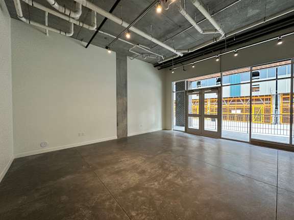 A large, empty room with a concrete floor and a view of a construction site through the windows at Paperbox Lofts Apartments, Salt Lake City