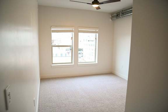 A room with a carpeted floor, a ceiling fan, and a window with a view of a building outside.