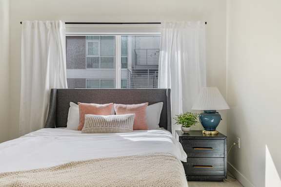 A bedroom with a large bed, a nightstand with a lamp, and a window with curtains. at Paperbox Lofts Apartments, Utah, 84101