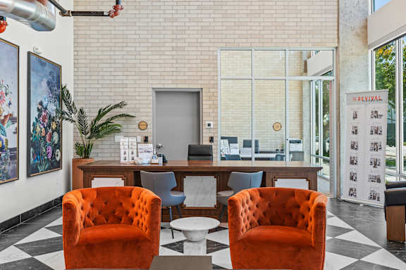 Two orange chairs are in front of a desk with a plant and pictures on the wall.