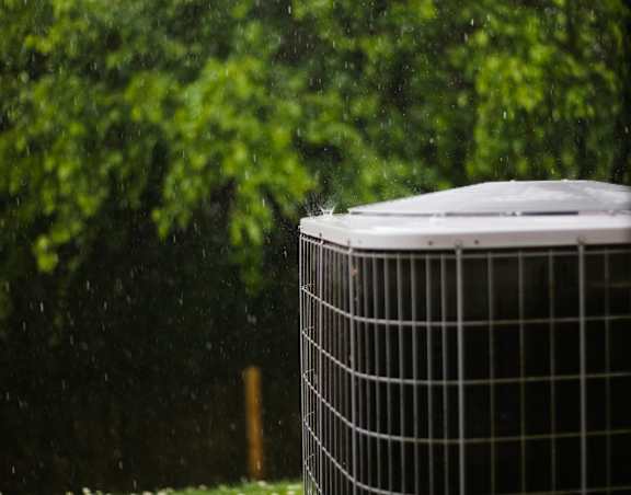 A black and white air conditioner unit is in front of a green bushy background.